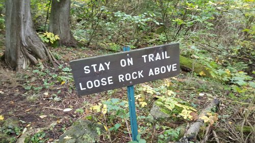 A wooden sign in a forest warns hikers to stay on the trail due to loose rocks above.