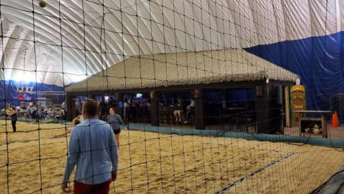 Indoor beach volleyball court with a thatched-roof bar and spectators in the background. Sand covers the floor.