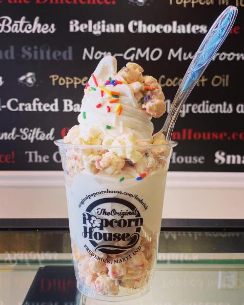 A clear cup filled with soft serve ice cream topped with colorful sprinkles and popcorn, against a branded background.