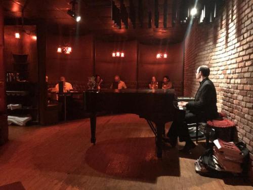 A pianist performs in a dimly lit bar, with patrons seated at tables enjoying the music.