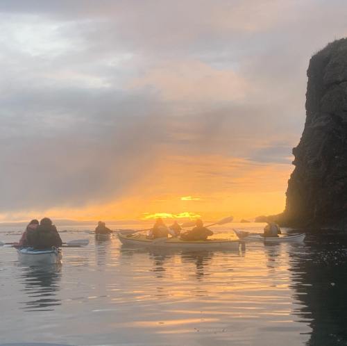 A group of kayakers paddles in calm waters at sunset, with a rocky cliff in the background and vibrant sky colors.