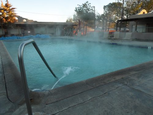 A steaming pool surrounded by a concrete deck and palm trees, with mist rising in the early morning light.