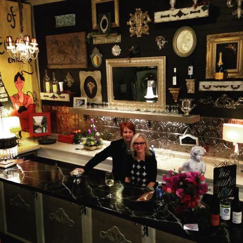 Two women pose at a stylish bar adorned with eclectic decor, flowers, and bottles, smiling for the camera.