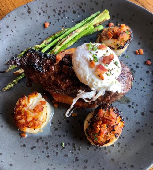 A plate featuring a grilled steak topped with a poached egg, asparagus, and two bacon-topped scallops.