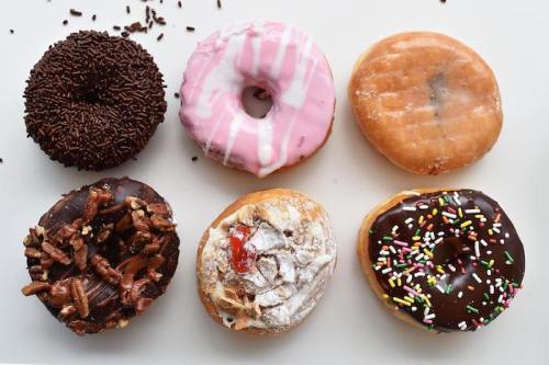 A variety of six donuts: chocolate sprinkles, pink icing, glazed, pecan, powdered sugar, and chocolate with colorful sprinkles.