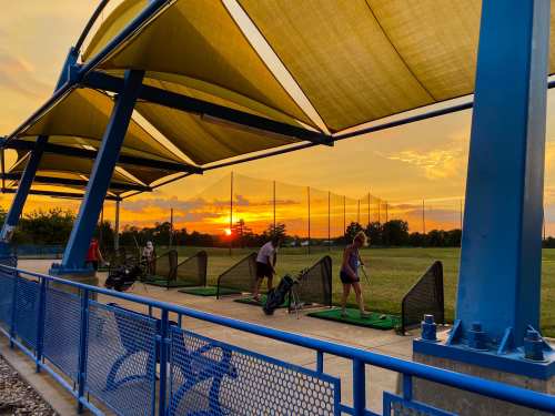 Golfers practice at a driving range under colorful sunset skies, with large canopies providing shade.