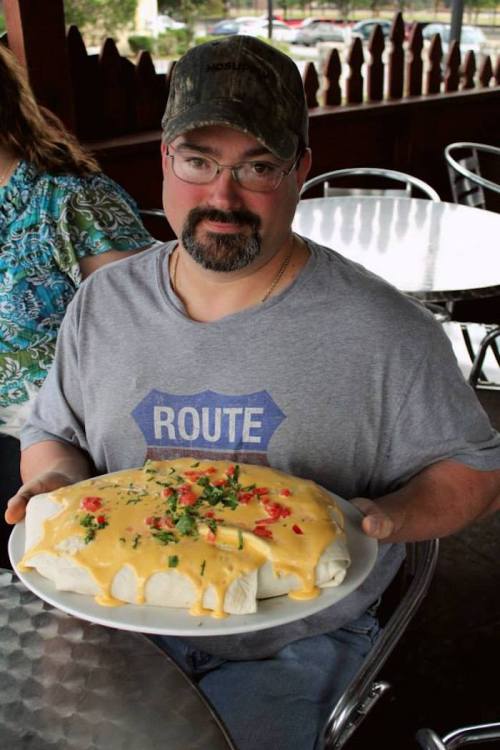 A man in a gray shirt holds a large plate of a burrito topped with cheese and colorful toppings at an outdoor restaurant.
