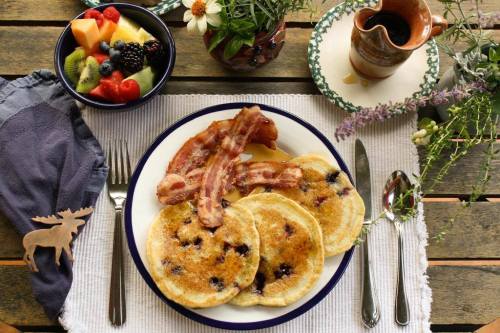 A plate of blueberry pancakes with bacon, surrounded by fruit salad and a small vase of flowers on a rustic table.