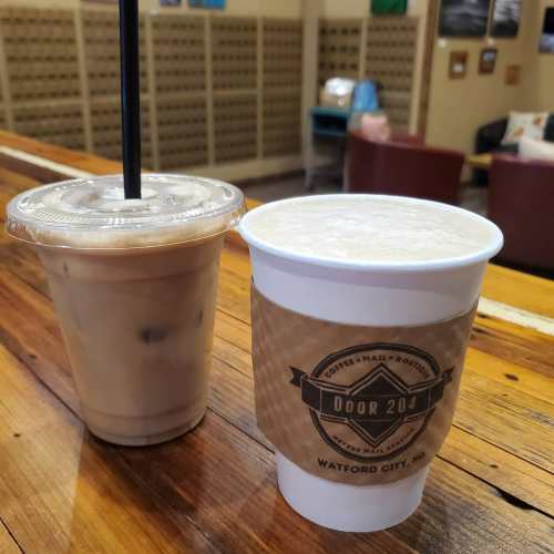 Two coffee drinks on a wooden table: one iced in a clear cup and one hot in a paper cup with a logo.