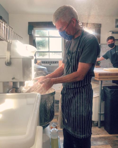 A person in an apron mixes ingredients in a kitchen, with flour dust in the air and another person in the background.