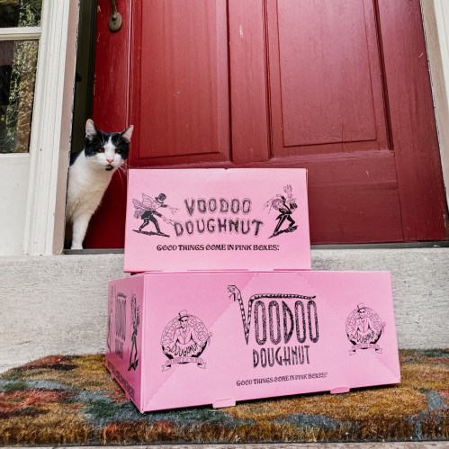 A cat peeks out from a doorway next to two pink Voodoo Doughnut boxes on a doorstep.