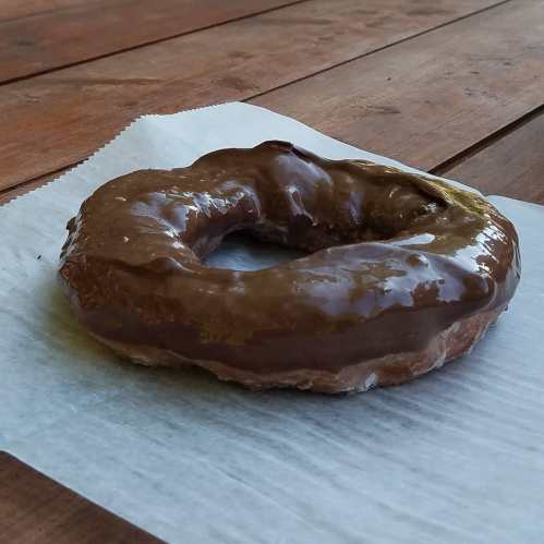 A chocolate-glazed donut resting on a piece of parchment paper on a wooden table.