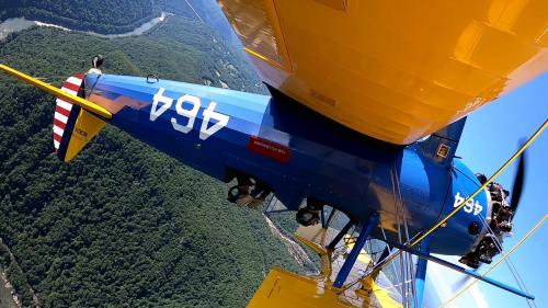 A blue and yellow biplane flying upside down over a lush green landscape with a river below.