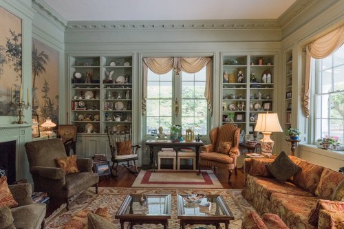 Cozy living room with green walls, shelves of collectibles, comfortable seating, and large windows letting in natural light.
