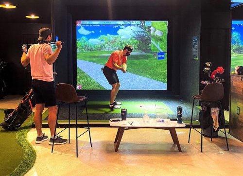 Two men in a golf simulator room; one is swinging a club while the other watches. A table with drinks is in the foreground.