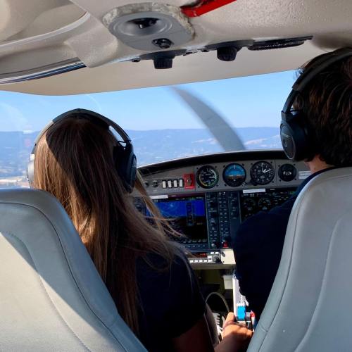 Two pilots in a small aircraft cockpit, wearing headsets, with instruments and a scenic view outside the window.