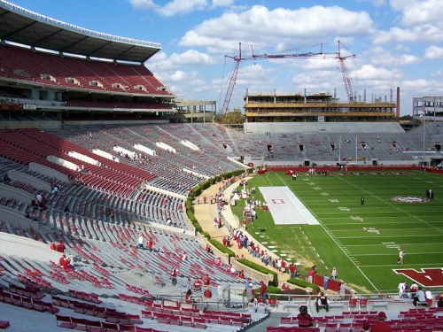 A view of a stadium with empty seats, a football field, and construction in the background under a blue sky.