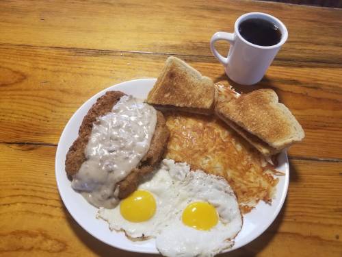 A plate with fried chicken, gravy, two sunny-side-up eggs, hash browns, and toast, alongside a cup of coffee.