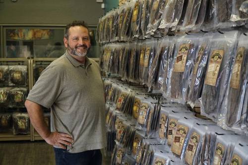 A man stands beside a wall of packaged jerky products in a store, smiling at the camera.