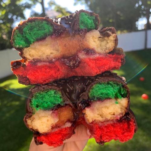 A hand holding a colorful layered pastry with green, red, and yellow sections, coated in chocolate and sprinkles.