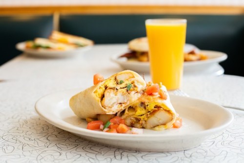 A close-up of a breakfast burrito filled with eggs and potatoes, topped with diced tomatoes, next to a glass of orange juice.
