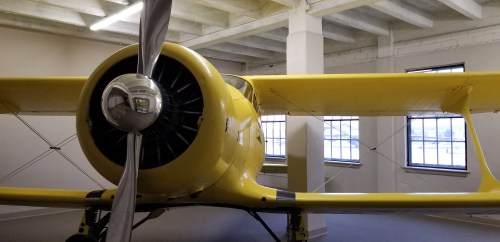 A yellow biplane with a large propeller displayed indoors, showcasing its wings and cockpit in a well-lit space.