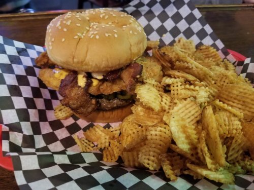 A sesame seed bun burger with bacon, pickles, and cheese, served with a side of wavy potato chips on a checkered paper.