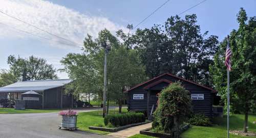 A small wooden building with a sign, surrounded by greenery, and an American flag in front on a sunny day.