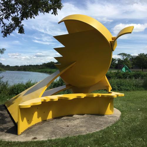 A large, abstract yellow sculpture resembling a bird, set against a scenic lakeside backdrop with green trees.