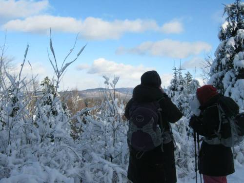 Two hikers in winter gear stand in a snowy landscape, gazing at a distant mountain under a blue sky.