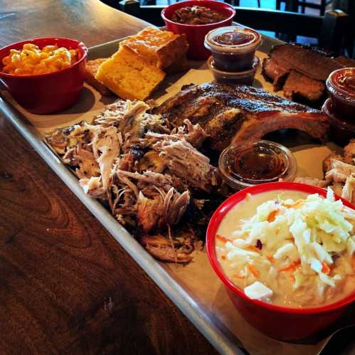 A platter of barbecue featuring pulled pork, ribs, cornbread, macaroni and cheese, coleslaw, and dipping sauces.