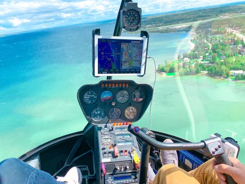 Cockpit view of a helicopter with navigation screens and controls, flying over a scenic blue lake and green landscape.