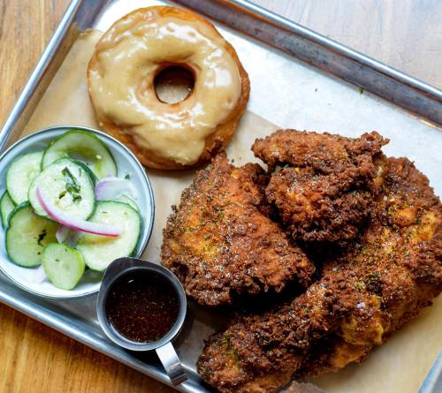 A metal tray with fried chicken, a glazed donut, and a side of pickled cucumbers with a small cup of sauce.