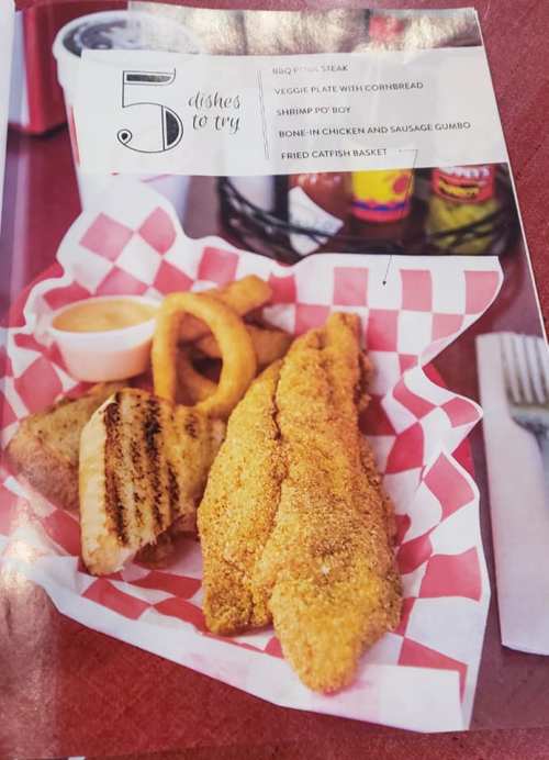 A plate featuring fried catfish, onion rings, and a side of dipping sauce on a red-checkered paper.