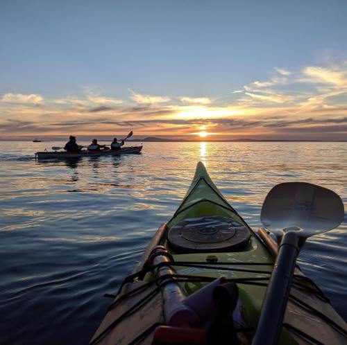 Two kayakers paddle in the distance as the sun sets over calm waters, viewed from the front of another kayak.