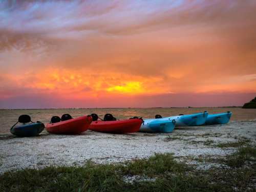 Five kayaks in red and blue lined up on a sandy shore, with a vibrant sunset and calm water in the background.