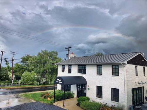 A white building with a black awning under a cloudy sky, featuring a rainbow in the background.