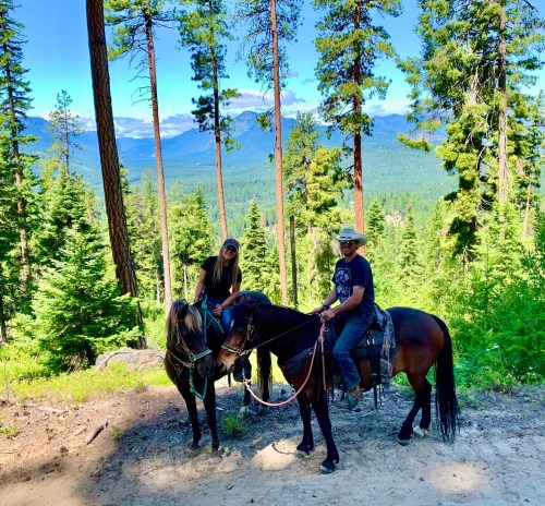 Two people on horseback in a forested area, surrounded by tall trees and mountains in the background.