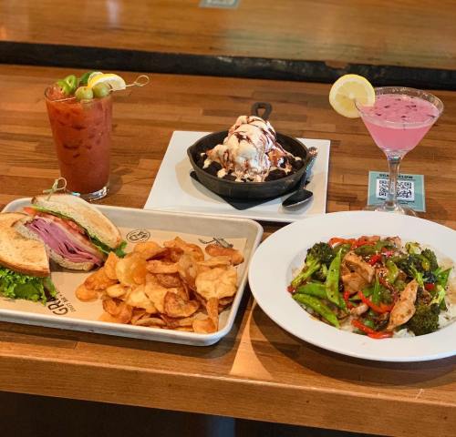A table with a sandwich, chips, stir-fried chicken and vegetables, ice cream dessert, and two colorful drinks.