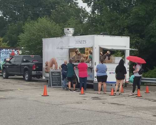 A food truck selling donuts with a line of people waiting, some holding umbrellas, in a parking lot.
