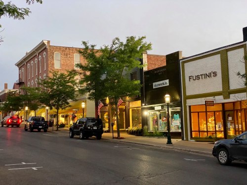 A charming street scene at dusk, featuring shops, trees, and parked cars, with American flags adorning the buildings.