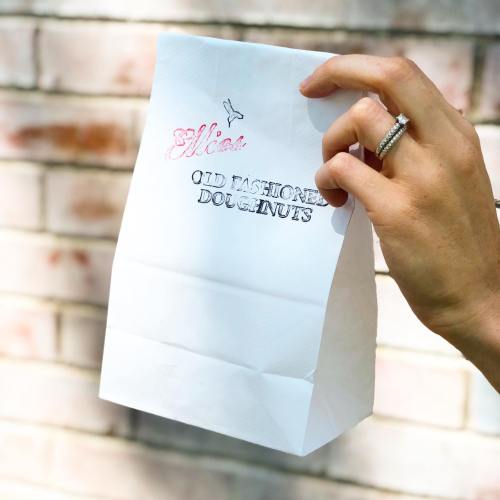 A hand holding a white paper bag with "Old Fashioned Doughnuts" printed on it against a brick wall.
