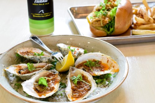 A plate of baked oysters with lemon and herbs, alongside a sandwich and fries, with a drink in the background.