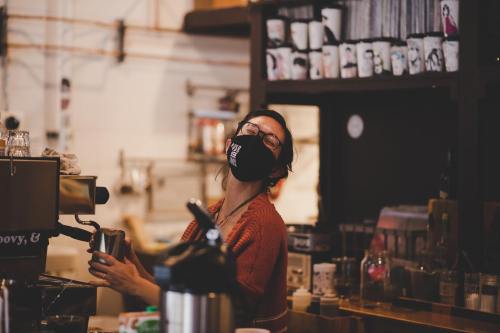 A barista in a mask smiles while preparing coffee in a cozy café filled with shelves of drinks and decor.