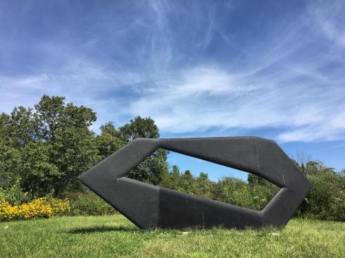 A large black sculpture shaped like an arrow, set against a blue sky and green landscape with trees and yellow flowers.