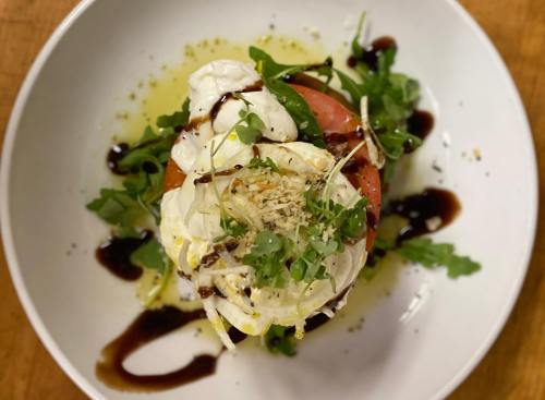 A fresh salad with sliced tomatoes, burrata cheese, greens, and a drizzle of balsamic reduction on a white plate.