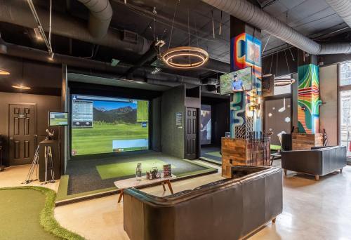 Indoor golf simulation area with a large screen, putting green, and modern seating. Brightly decorated walls.