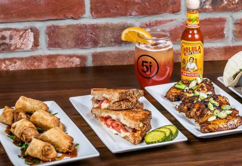 A table with three plates of food: spring rolls, a grilled sandwich, and marinated chicken, alongside a drink and hot sauce.