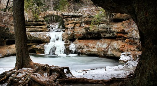 A serene winter scene featuring a frozen waterfall and a stone bridge surrounded by snow-covered rocks and trees.