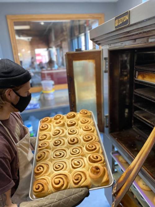 A person wearing a mask holds a tray of freshly baked cinnamon rolls, ready to be taken out of the oven.